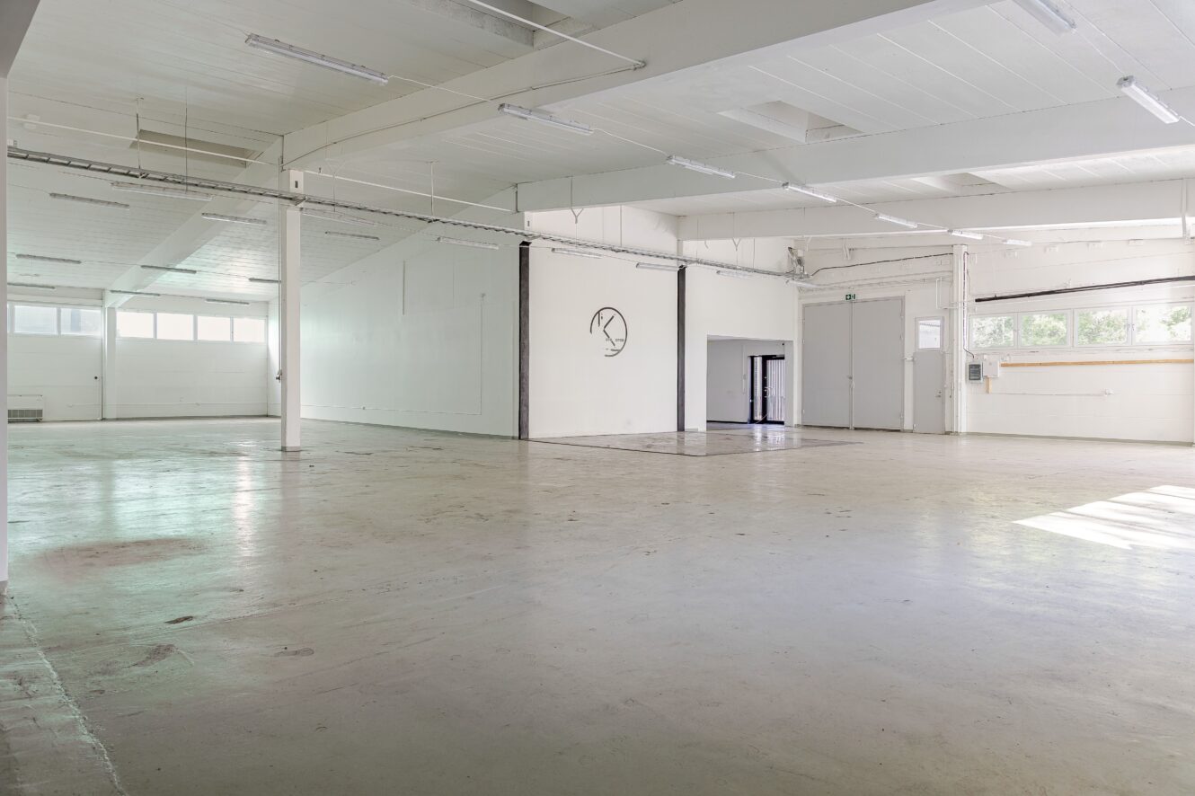 Rymlig, tom industrihall med vita väggar, stora fönster och lysrörsbelysning.