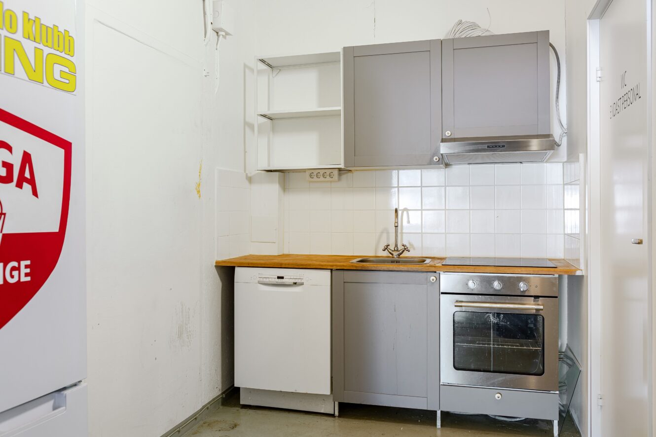 Litet kök med grå skåpluckor, bänkskiva i trä, inbyggd ugn, spishäll, diskmaskin och diskho mot en vit kaklad vägg. Ett kylskåp är delvis synligt till vänster.