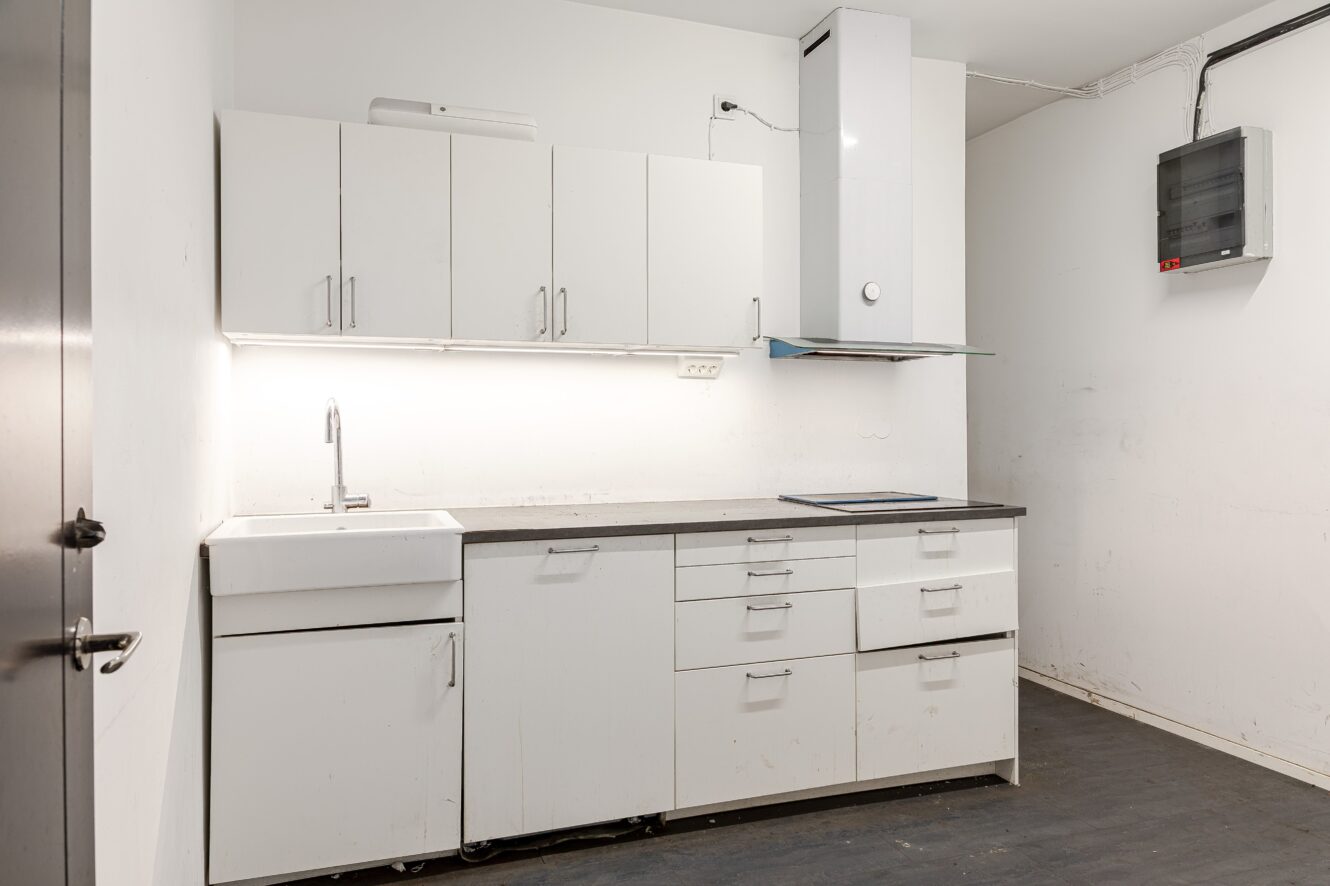 Ett modernt, minimalistiskt kök med vita skåp, diskho, elektrisk spishäll, väggmonterade skåp och belysning under skåpen.