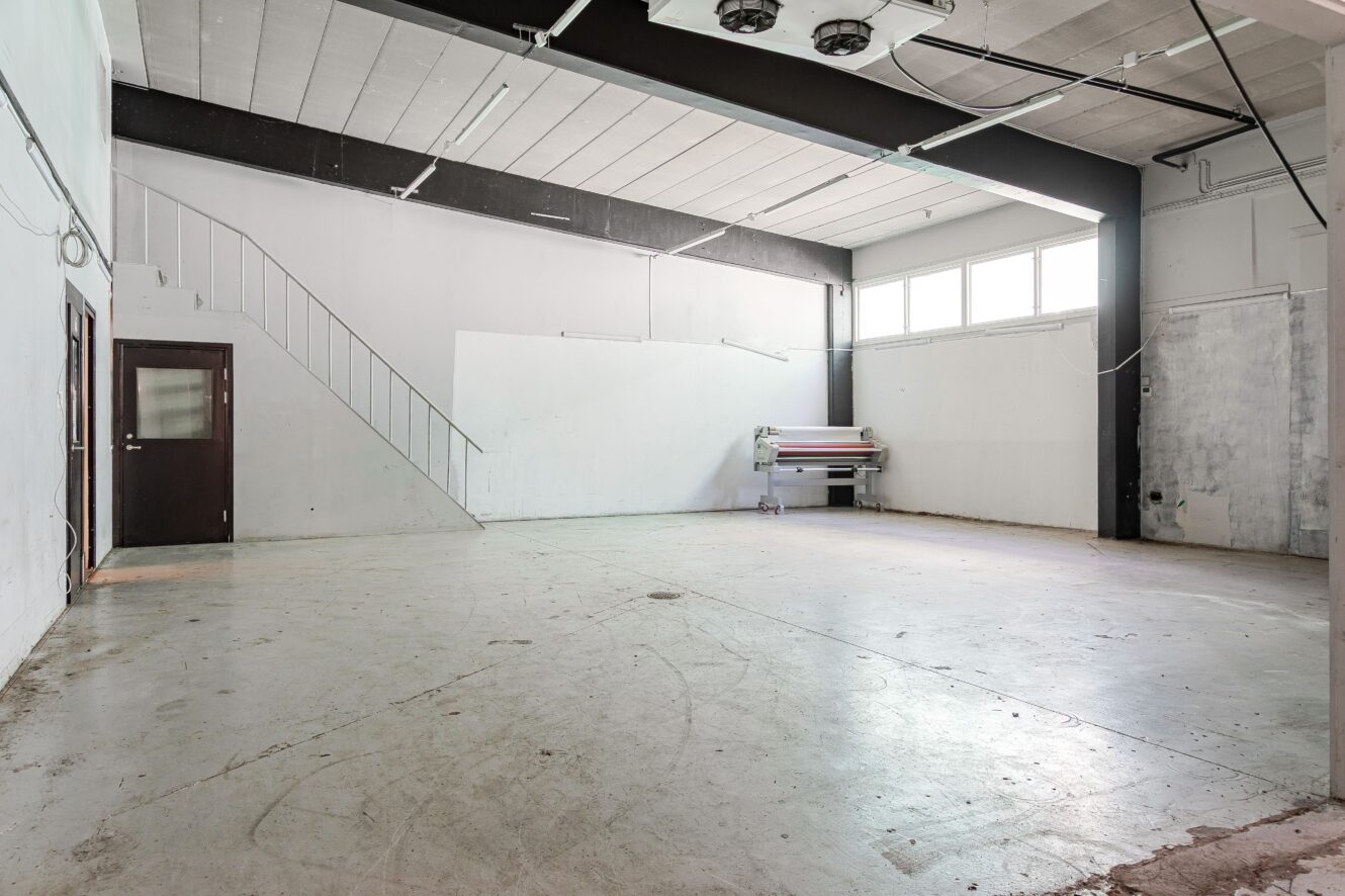 Rymligt, tomt industrirum med betonggolv, vita väggar, en trappa, en mörk dörr och stora fönster som släpper in naturligt ljus. En stor skrivare står mot den bortre väggen.