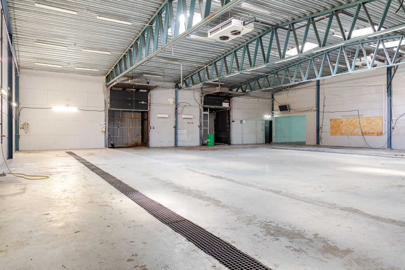 Stort, tomt industrilager med betonggolv, synliga stålbalkar, högt i tak och flera stora lastportar längs bakväggen.