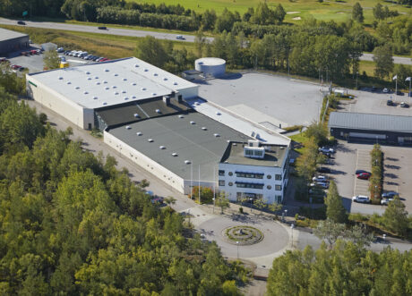 Flygfoto över en stor industribyggnad med en intilliggande parkeringsplats, omgiven av träd och kantad av en väg.