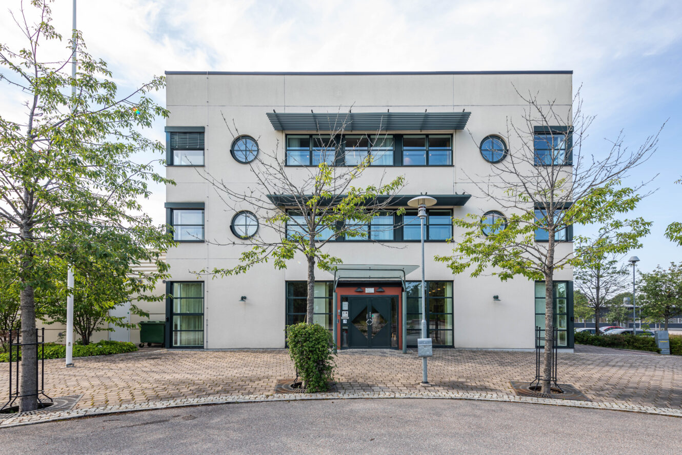 En modern kontorsbyggnad i tre våningar med stora fönster, en entrédörr i glas och en asfalterad uppfart kantad av unga träd.