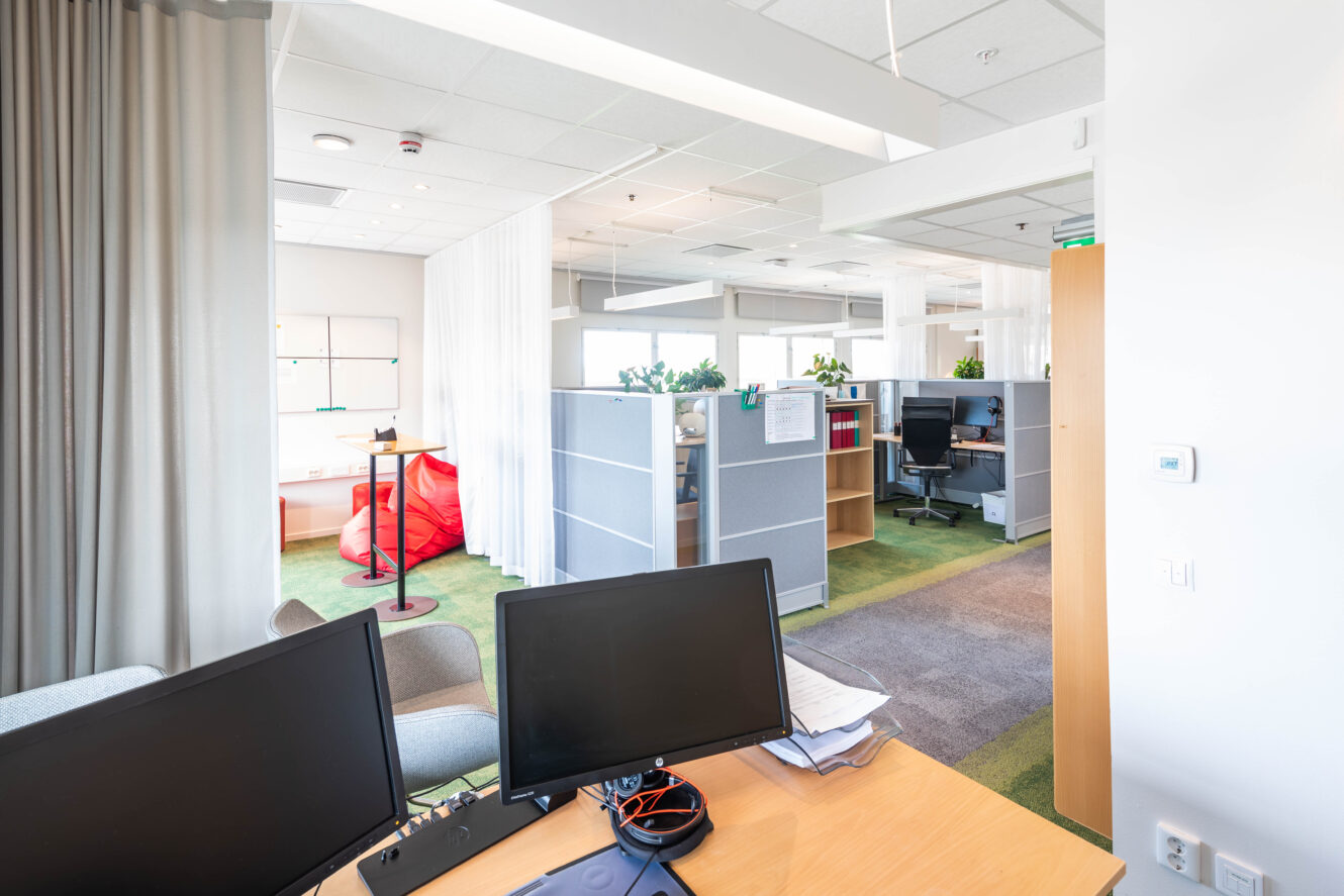 Modernt öppet kontorslandskap med bås, skrivbord, stolar, datorer och grön matta. Naturligt ljus kommer in genom stora fönster. Röda stolar med saccosäckar syns i bakgrunden.