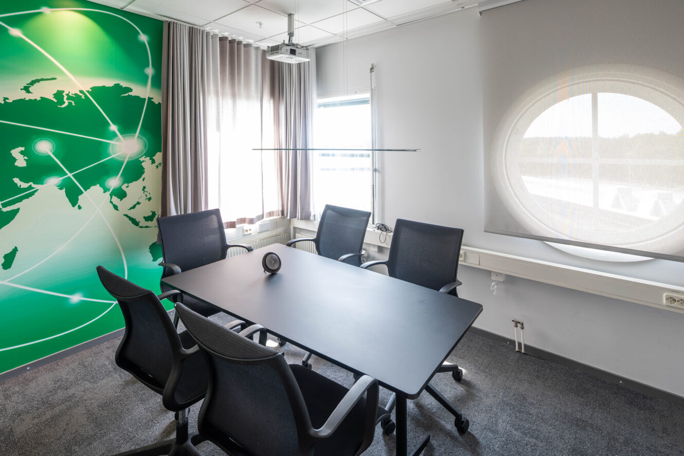 Ett modernt konferensrum med fyra svarta stolar runt ett rektangulärt bord, stora fönster och en väggmålning med en grön världskarta och nätverkslinjer.