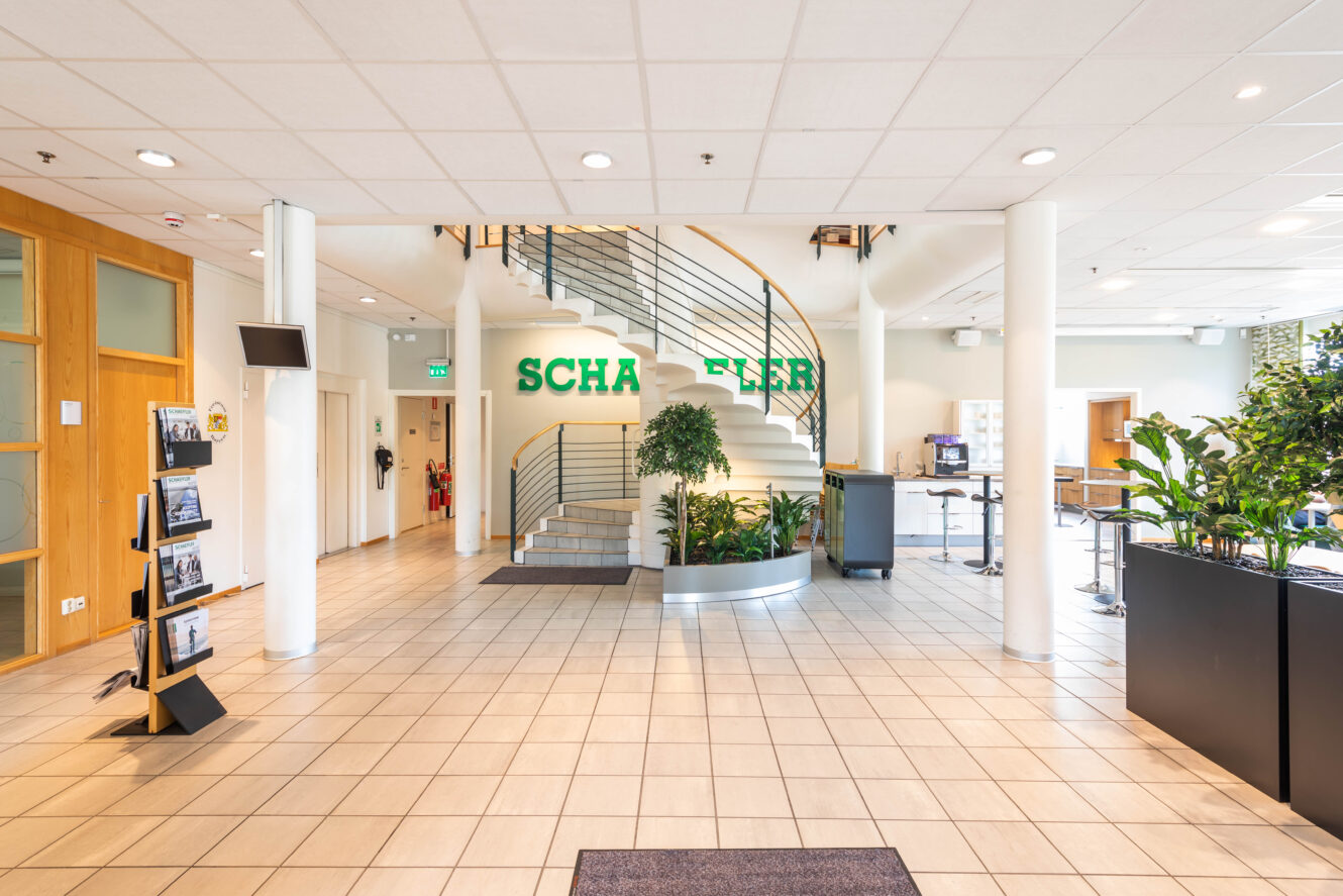 Rymlig lobby med klinkergolv, trappa med gröna bokstäver SCHA och FLER på väggen, växter, broschyrställ och sittgrupp till höger.