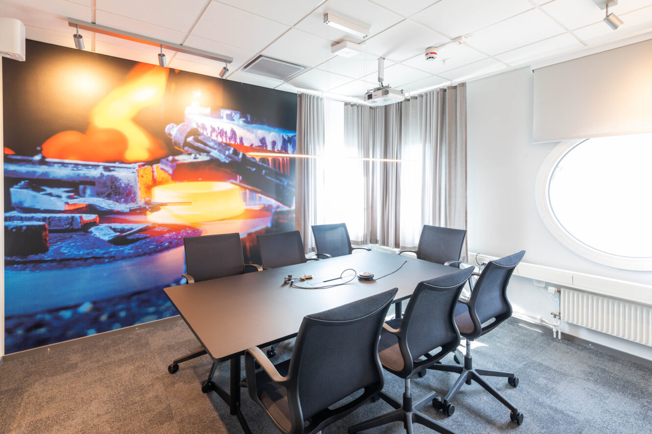Modernt konferensrum med sex stolar runt ett rektangulärt bord, stor väggkonst av industriellt metallarbete och ett runt fönster som släpper in naturligt ljus.