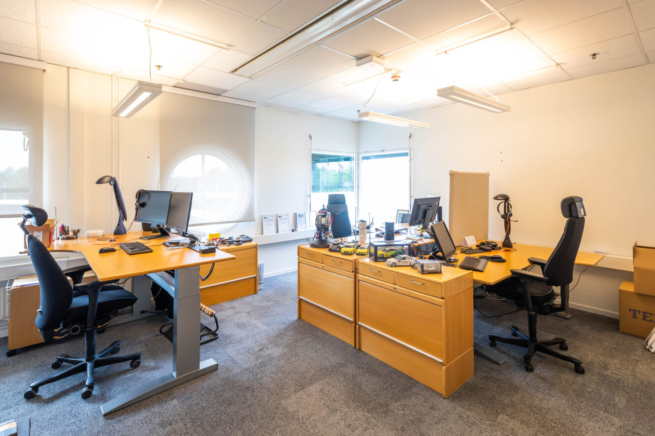 Ett modernt kontor med fyra skrivbord, datorer, kontorsstolar, utspridda papper och lådor, under starka taklampor och stora fönster.