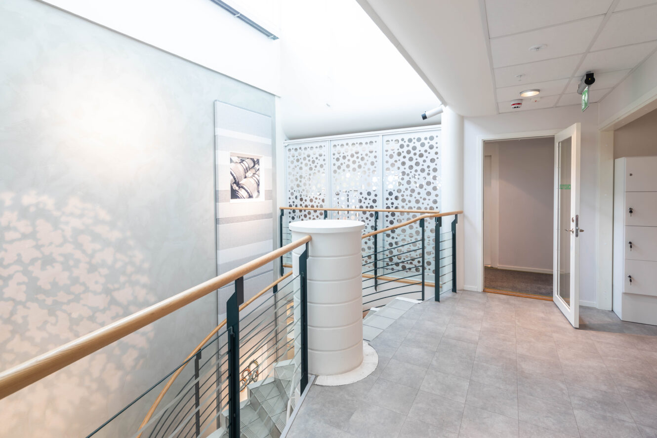 En modern, ljus hall med grått klinkergolv, räcke i metall och trä, skåp, öppna dörrar och dekorativa väggpaneler med runda utskärningar.