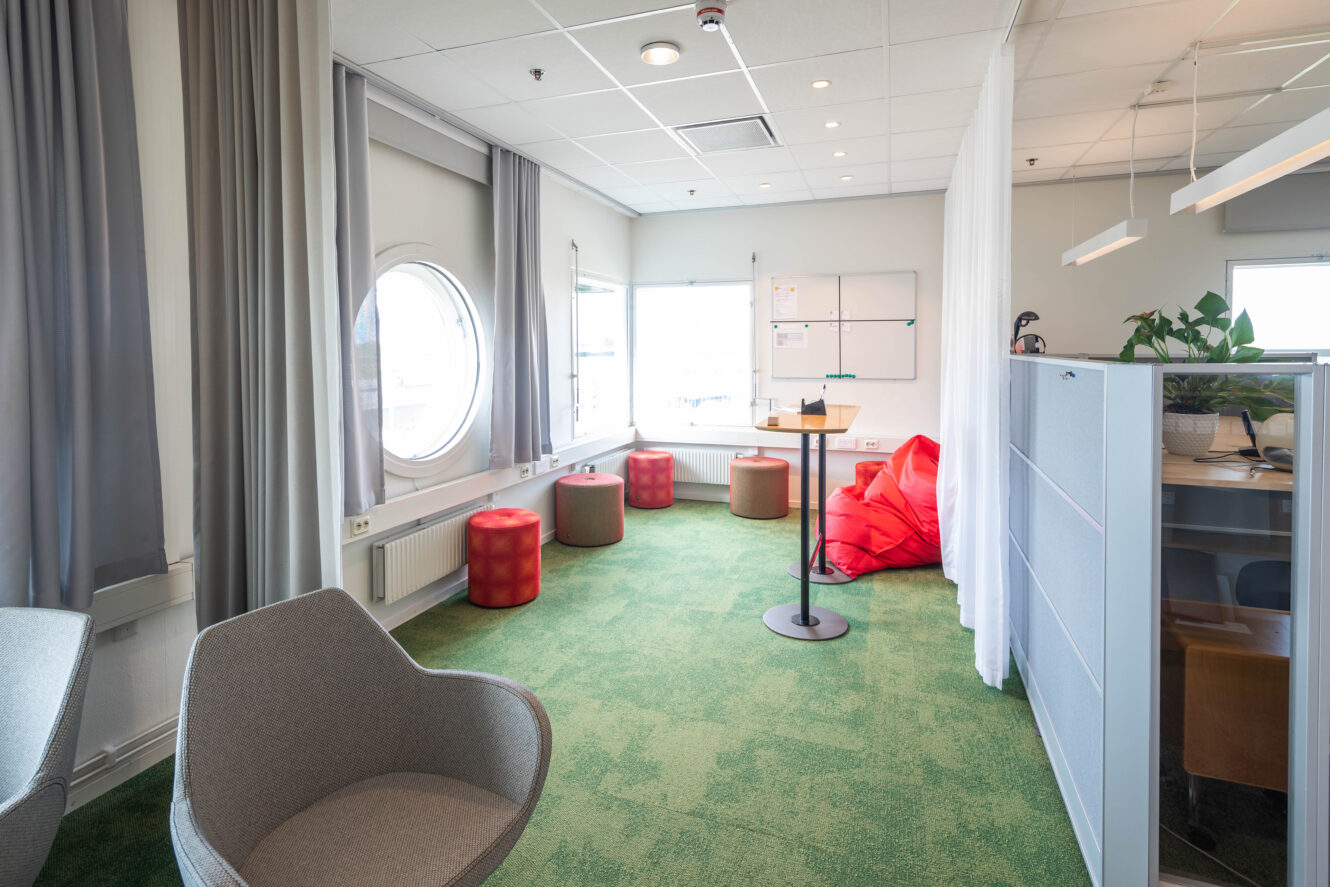 Modernt kontorsrum med grön matta, grå stolar, röda ottomaner, sittsäckar, runt fönster, whiteboard och avgränsad arbetsyta med en växt.