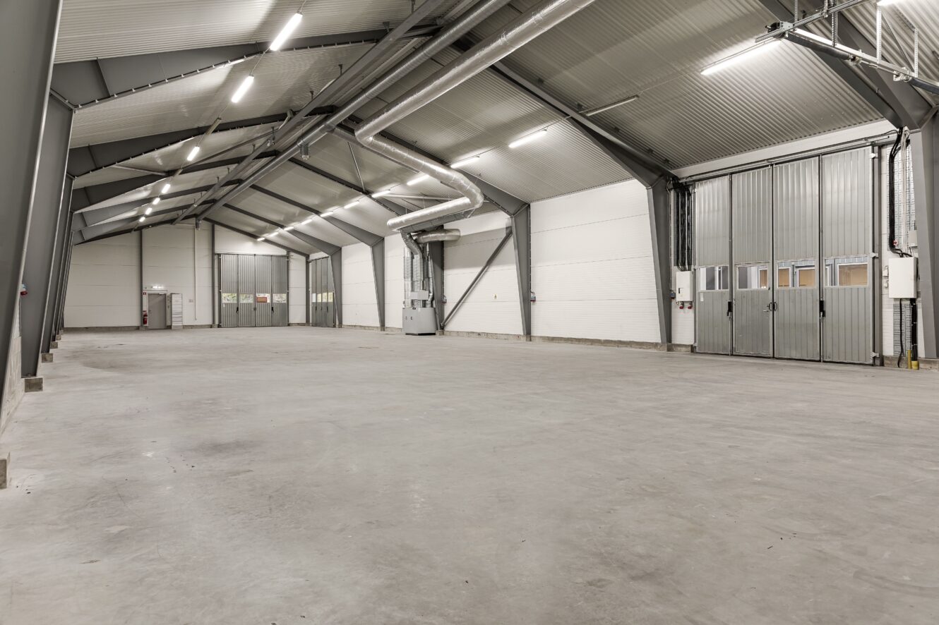 Stort tomt industrilager med betonggolv, vita väggar, högt i tak, metallbalkar och breda stängda dörrar till höger.