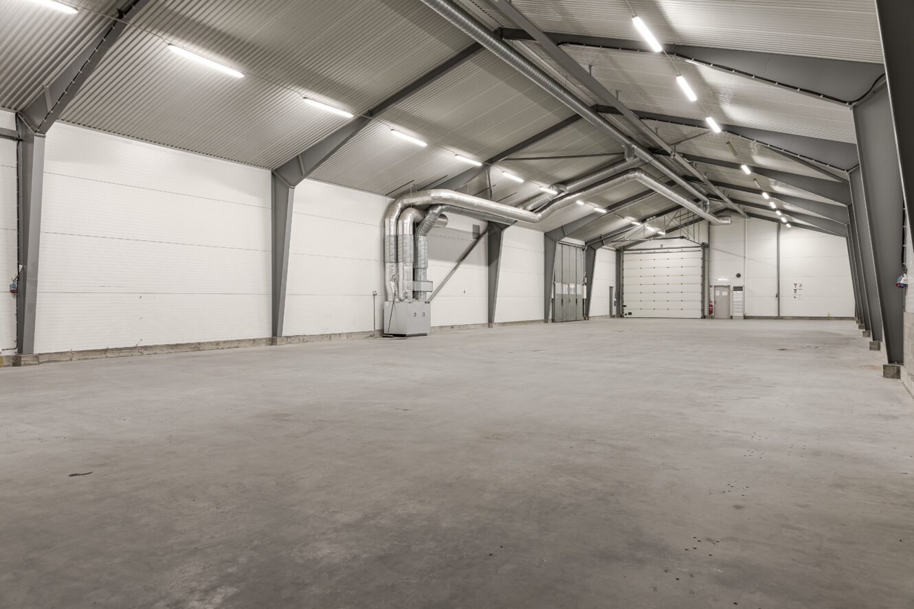 Tomt industrilager med vita väggar, gråa stålbalkar, takbelysning, synliga ventilationskanaler och en stor stängd garageport längst bort.