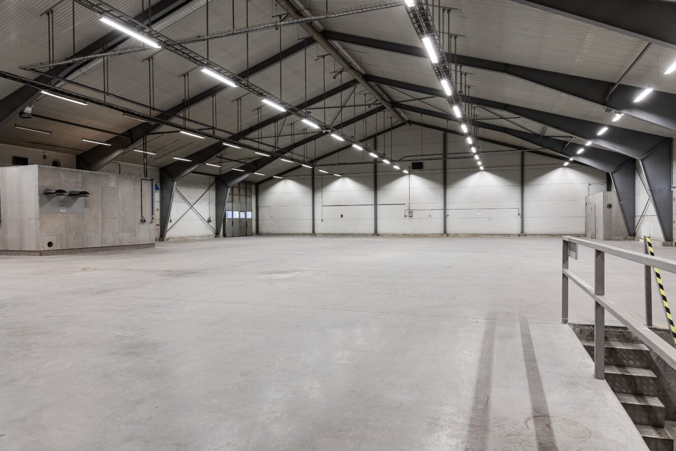 Stor, tom industrilagerlokal med högt i tak, betonggolv, metallbalkar, takbelysning och en upphöjd plattform med säkerhetsräcke till höger.