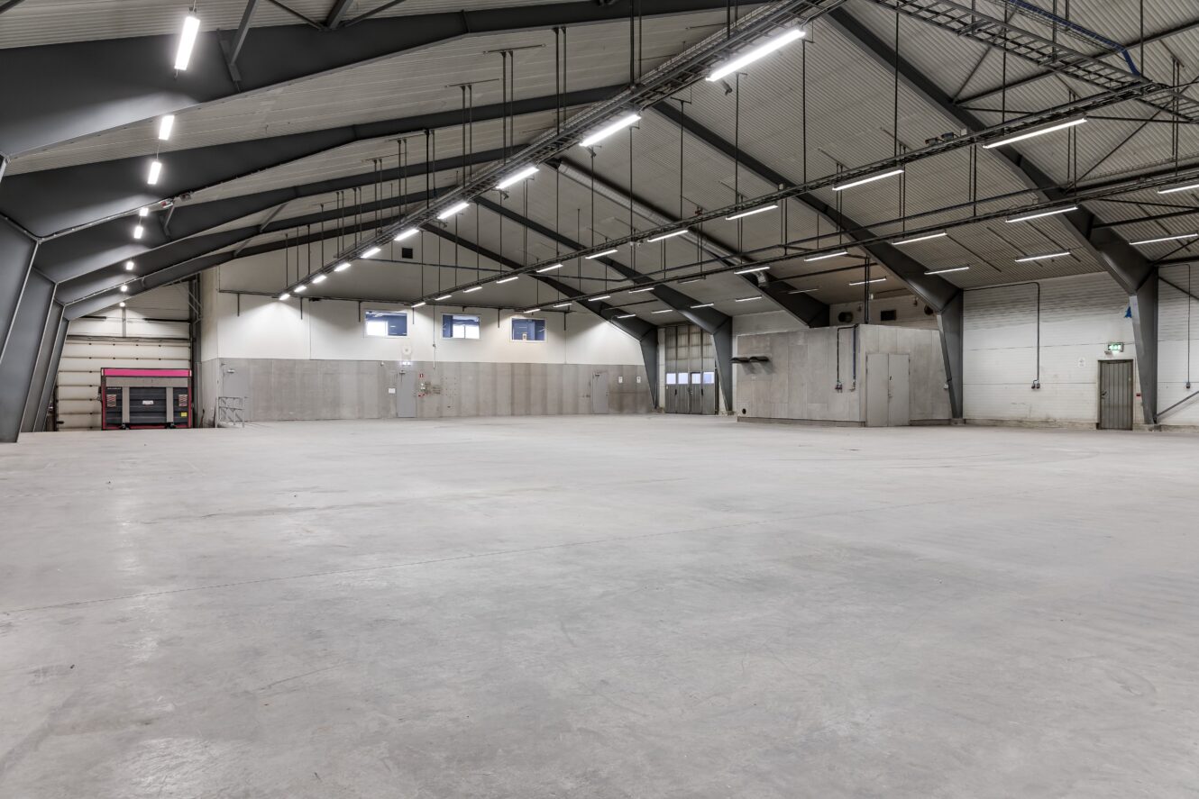 Stort tomt industrilager med betonggolv, högt i tak, synliga bjälkar och lysrörsbelysning. Några fönster och dörrar är synliga längs väggarna.