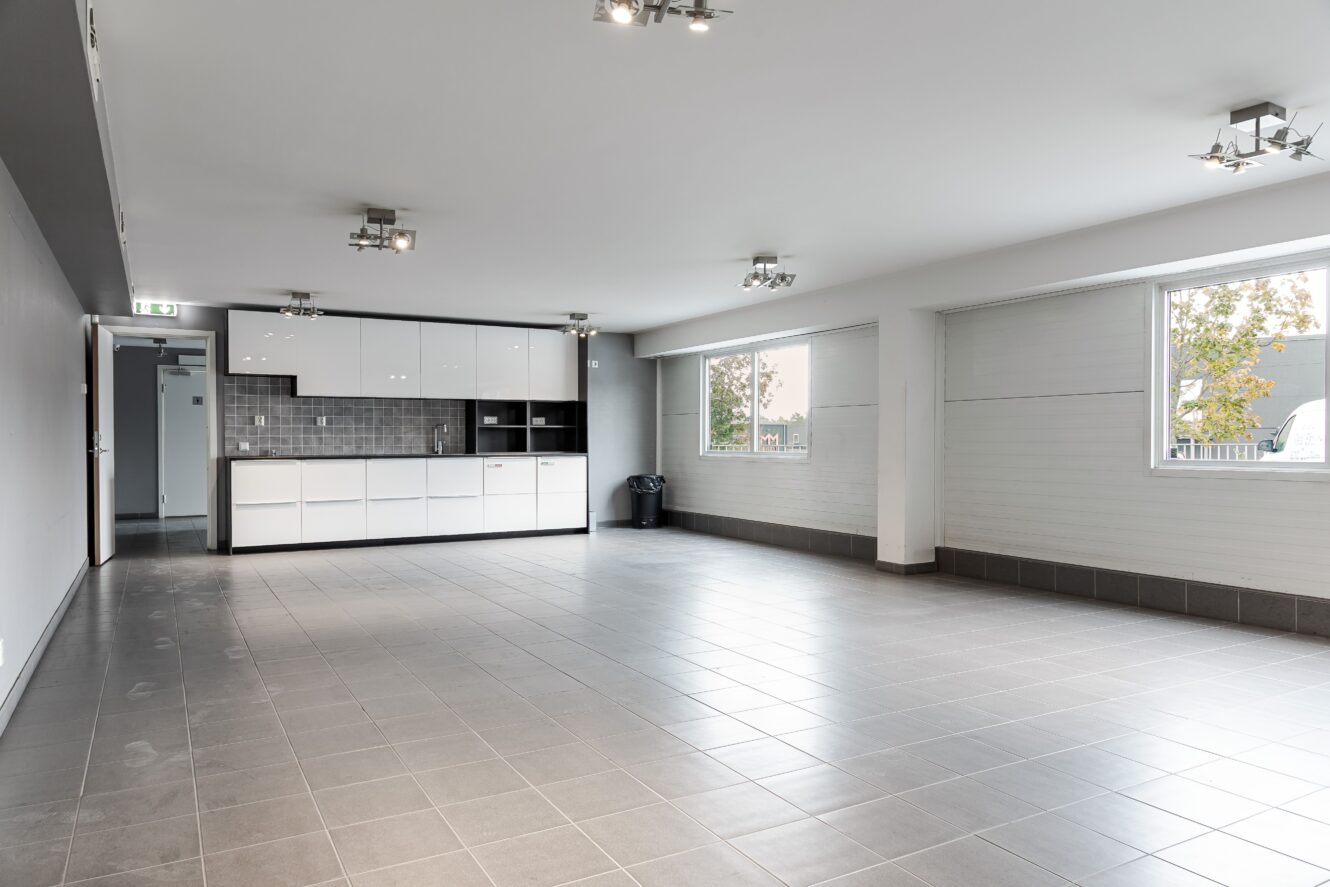 Rymligt, tomt rum med klinkergolv, vita skåp längs bakväggen, grått stänkskydd, stora fönster och taklampor.
