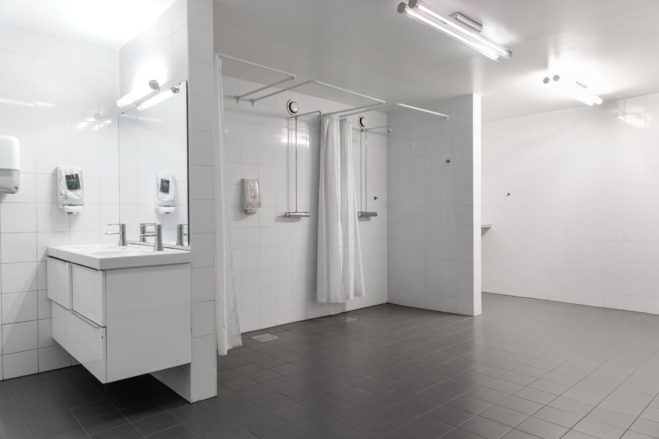 Ett modernt offentligt duschrum med vitkaklade väggar, grått klinkergolv, två handfat med speglar och två duschkabiner åtskilda av vita draperier.