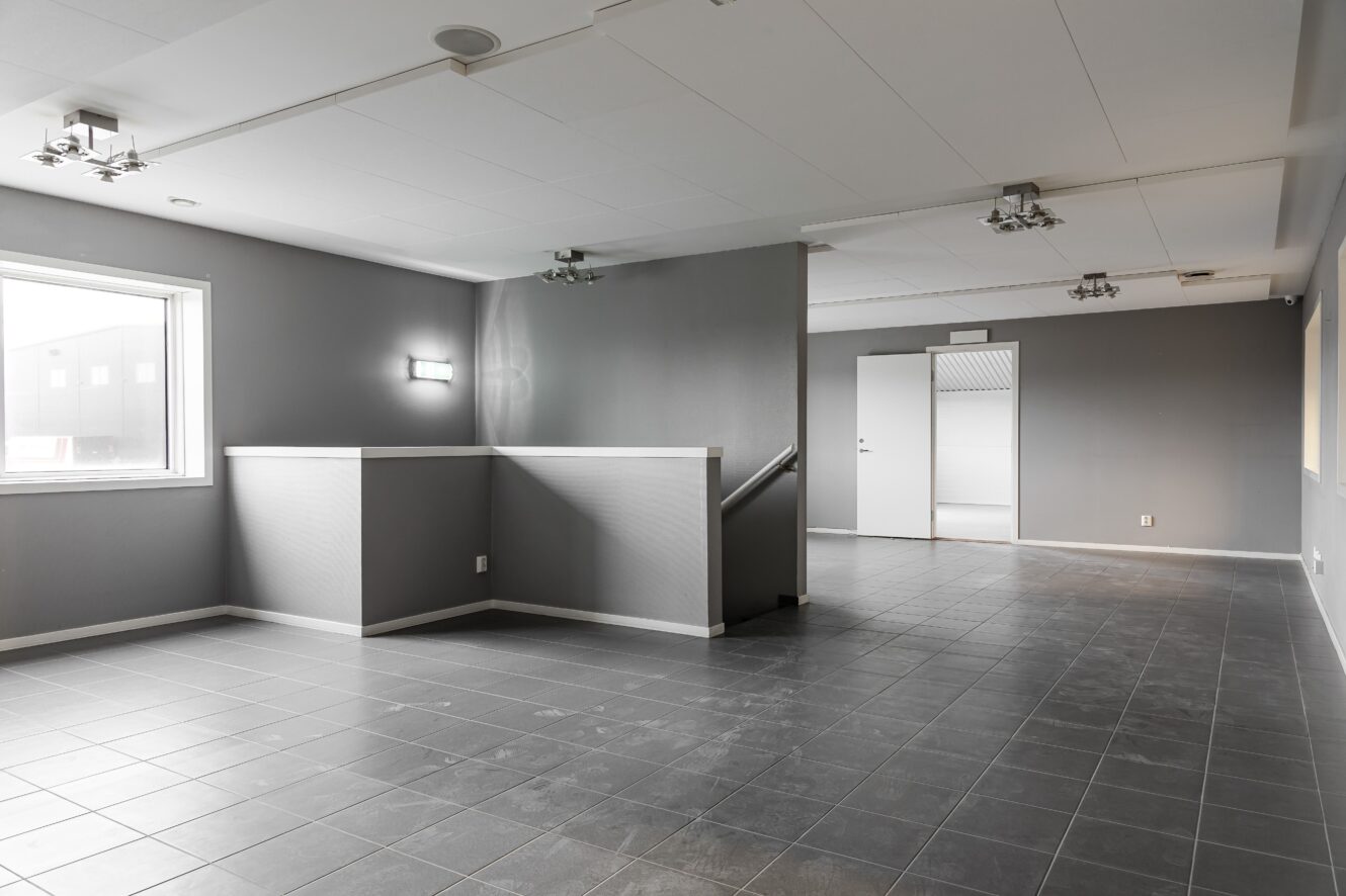 Modernt tomt rum med grått klinkergolv, grå väggar, ett fönster till vänster, väggmonterade lampor och en trappa med en del av väggen. Vit dörr och hall syns i bakgrunden.