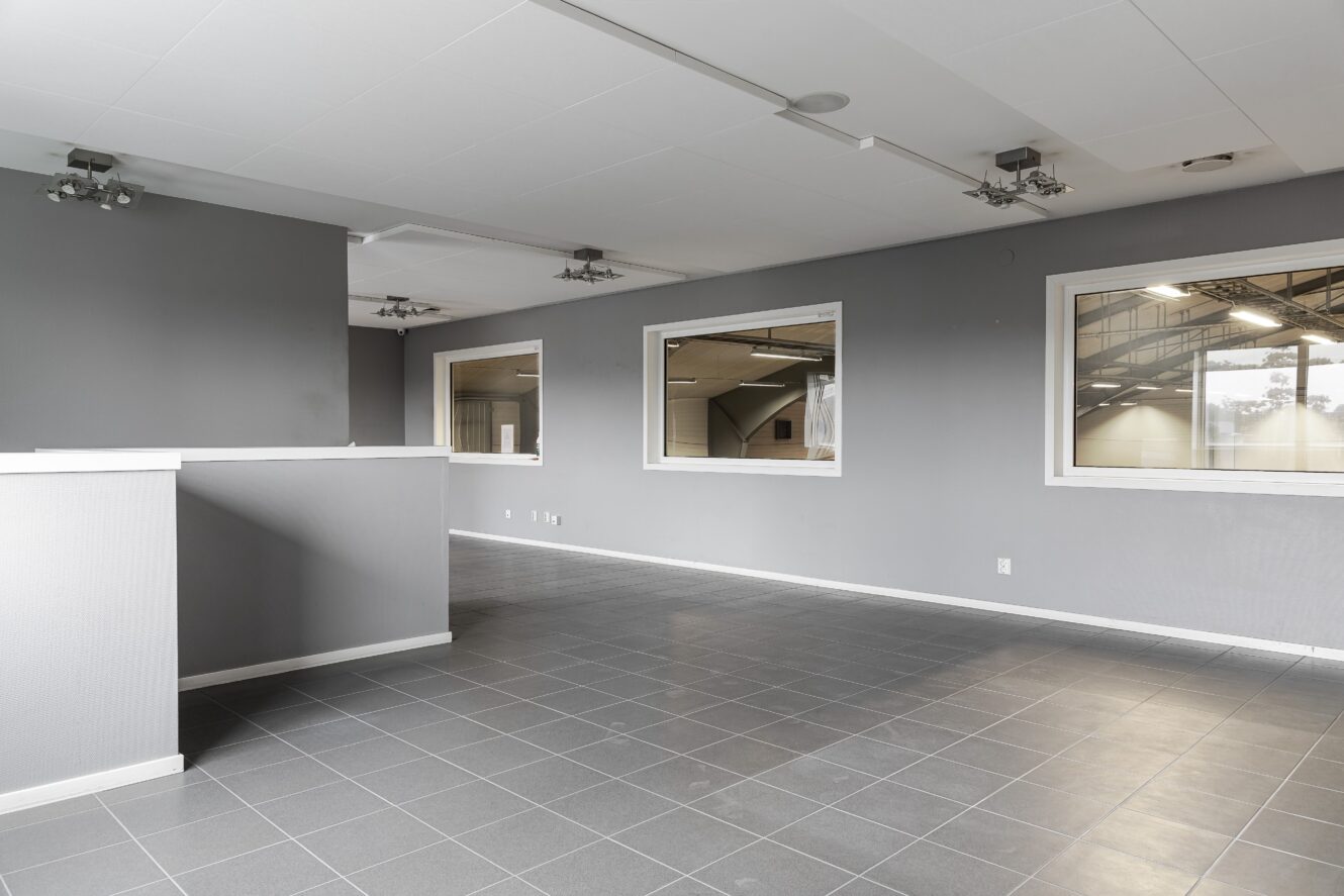 Tomt modernt rum med grått klinkergolv, grå väggar och stora innerfönster med utsikt över en intilliggande hall. Taklampor och vita lister är synliga.