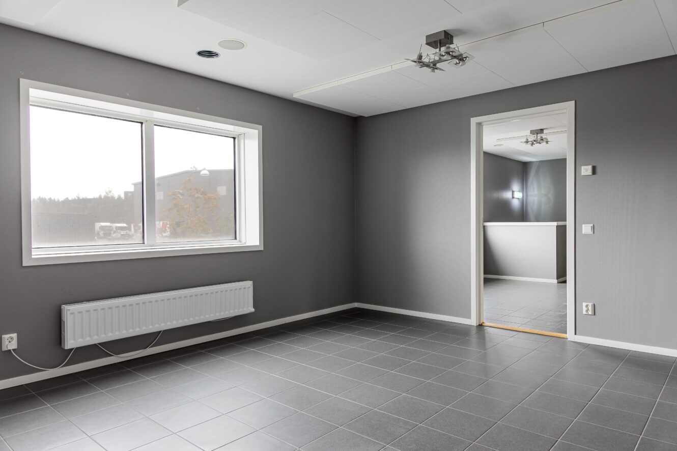 Tomt rum med grått klinkergolv, grå väggar, ett stort fönster, vitt tak, taklampa och en öppen dörr till ett annat liknande rum.