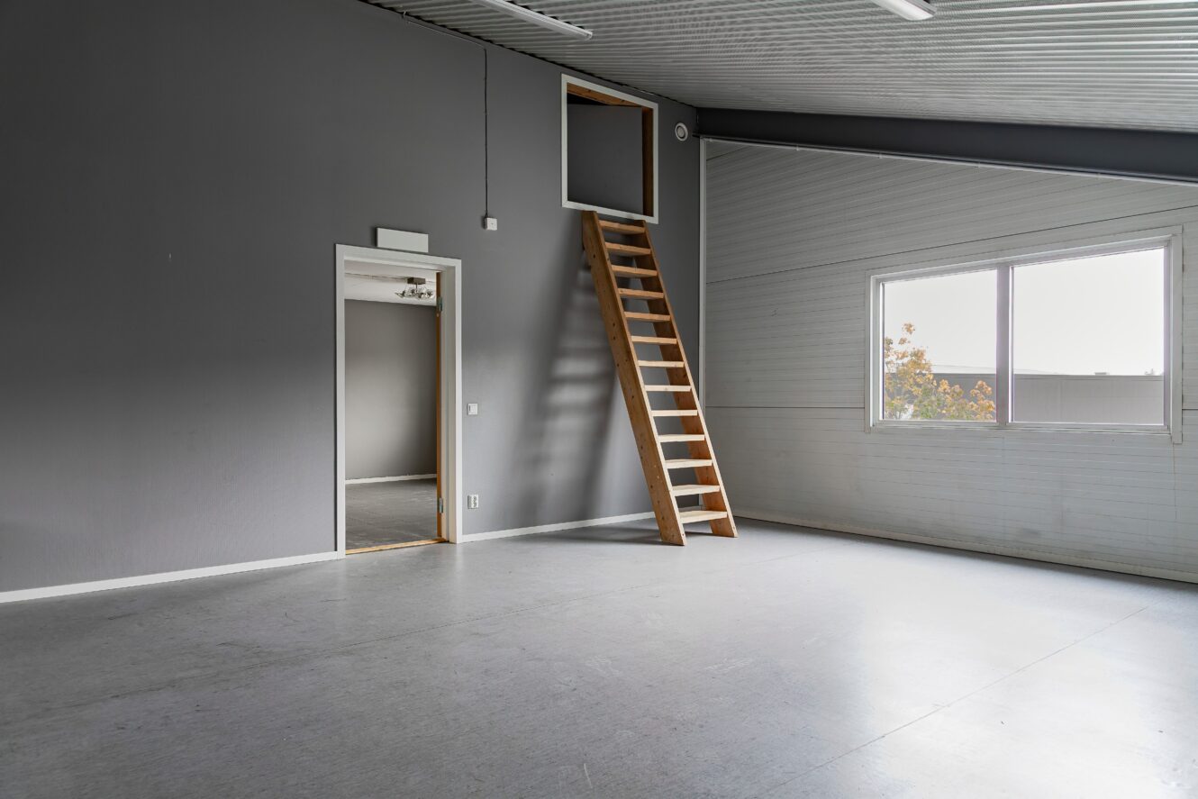 Tomt industrirum med grå väggar, stort fönster, öppen dörröppning till ett annat rum och trästege som leder till ett loft eller en övre öppning.