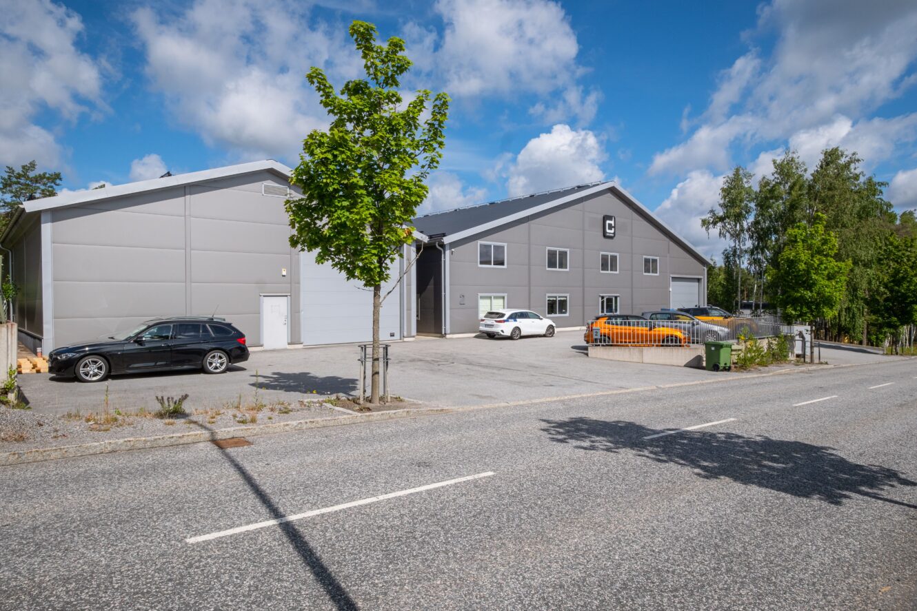 Ett grått industrilager med flera parkerade bilar framför, kantat av träd och en väg under en delvis molnig himmel.