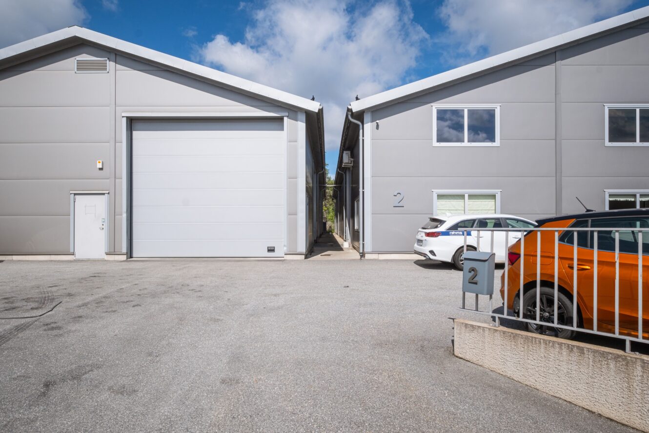 Två gråa industribyggnader med numrerade skyltar skiljs åt av en uppfart; bilar står parkerade framför och himlen är delvis molnig.