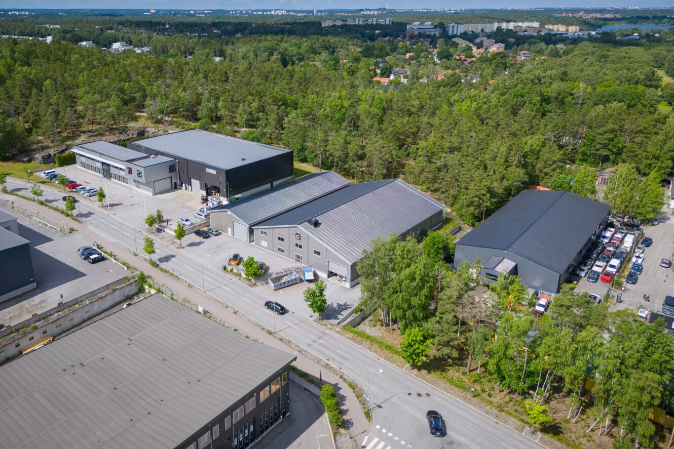Flygbild över ett industriområde med flera stora lagerbyggnader omgivna av träd och några parkerade bilar längs vägen.