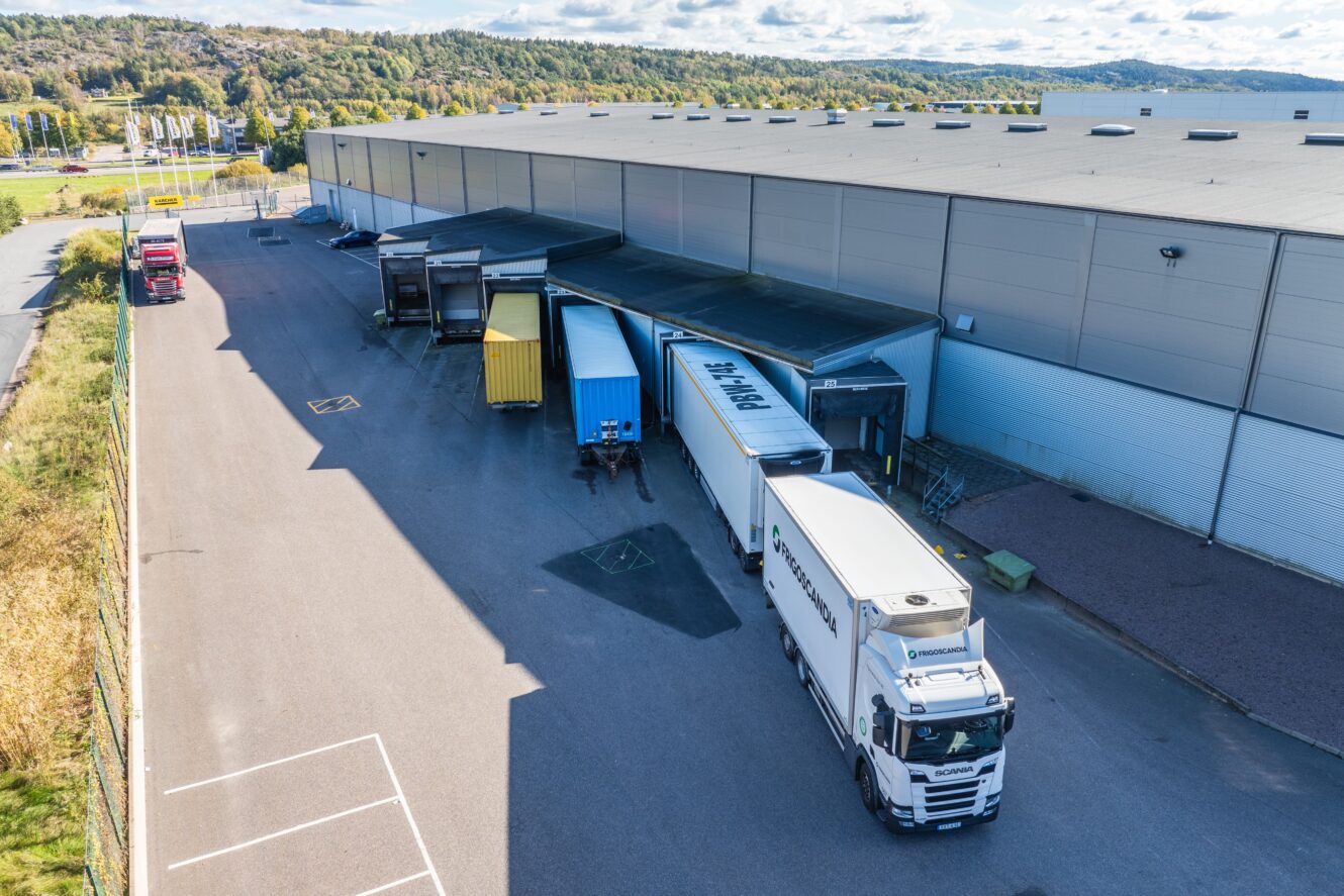 Flygfoto över en lastkaj med flera lastbilar parkerade som lossar eller lastar varor; en stor byggnad och träd syns i bakgrunden.