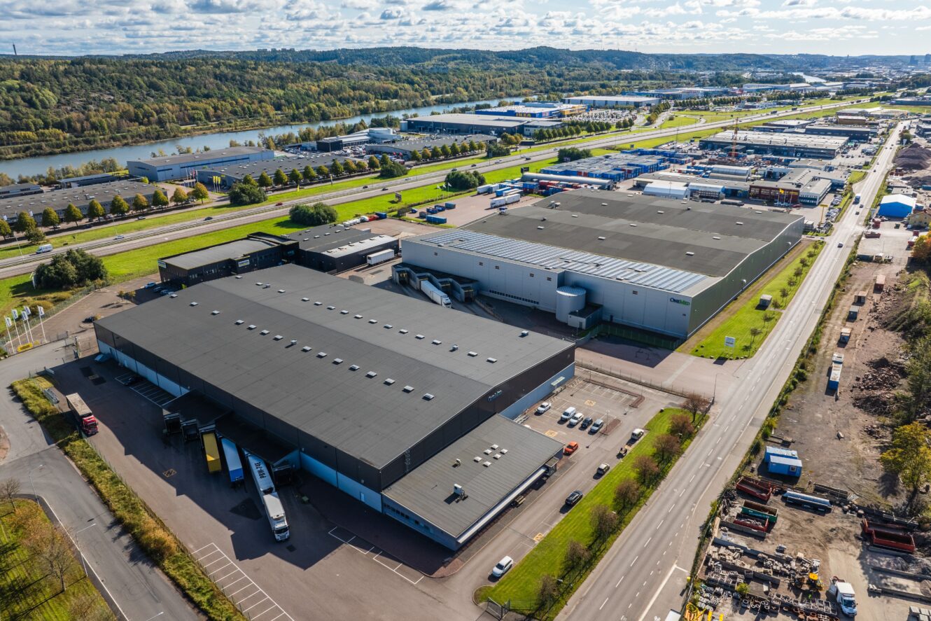 Flygfoto över industrilager med lastkajer, lastbilar och intilliggande vägar; grönt landskap och en flod i bakgrunden.