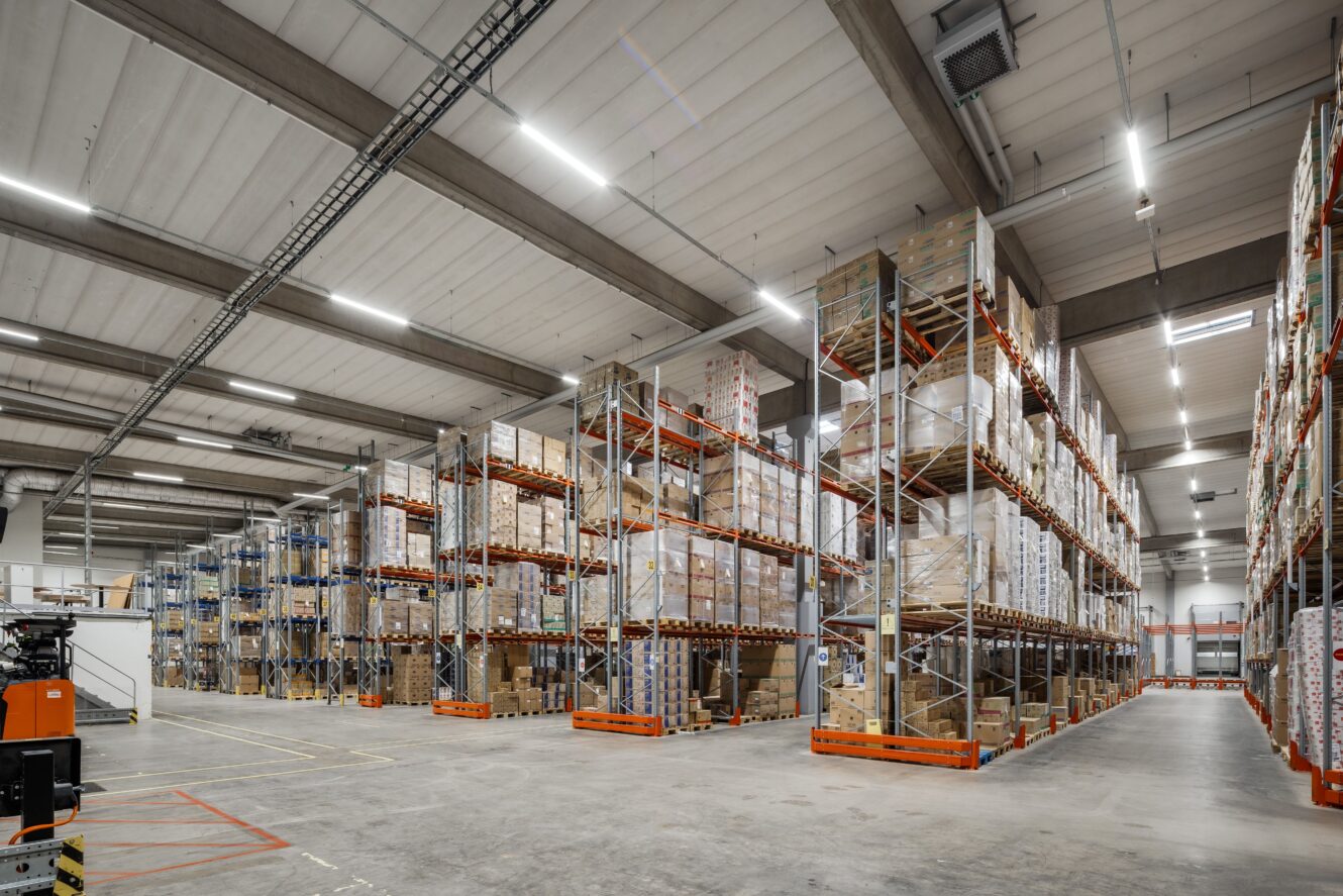 Stor lagerlokal med höga hyllor fulla av lådor och varor, breda gångar och stark takbelysning.
