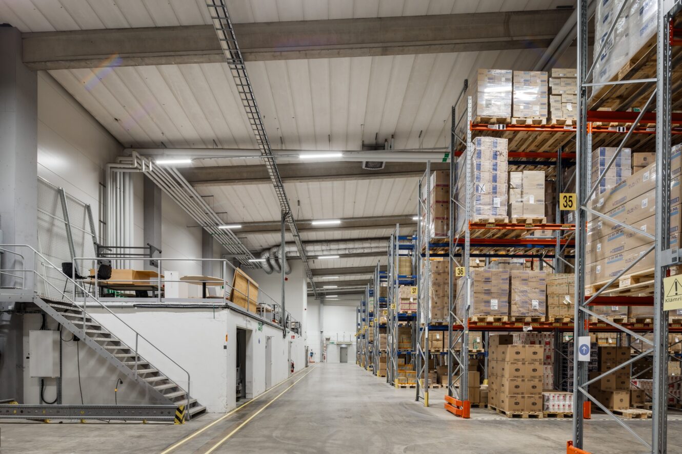 Stor lagerlokal med höga hyllor staplade med lådor och pallar, betonggolv och metalltrappa på vänster sida.