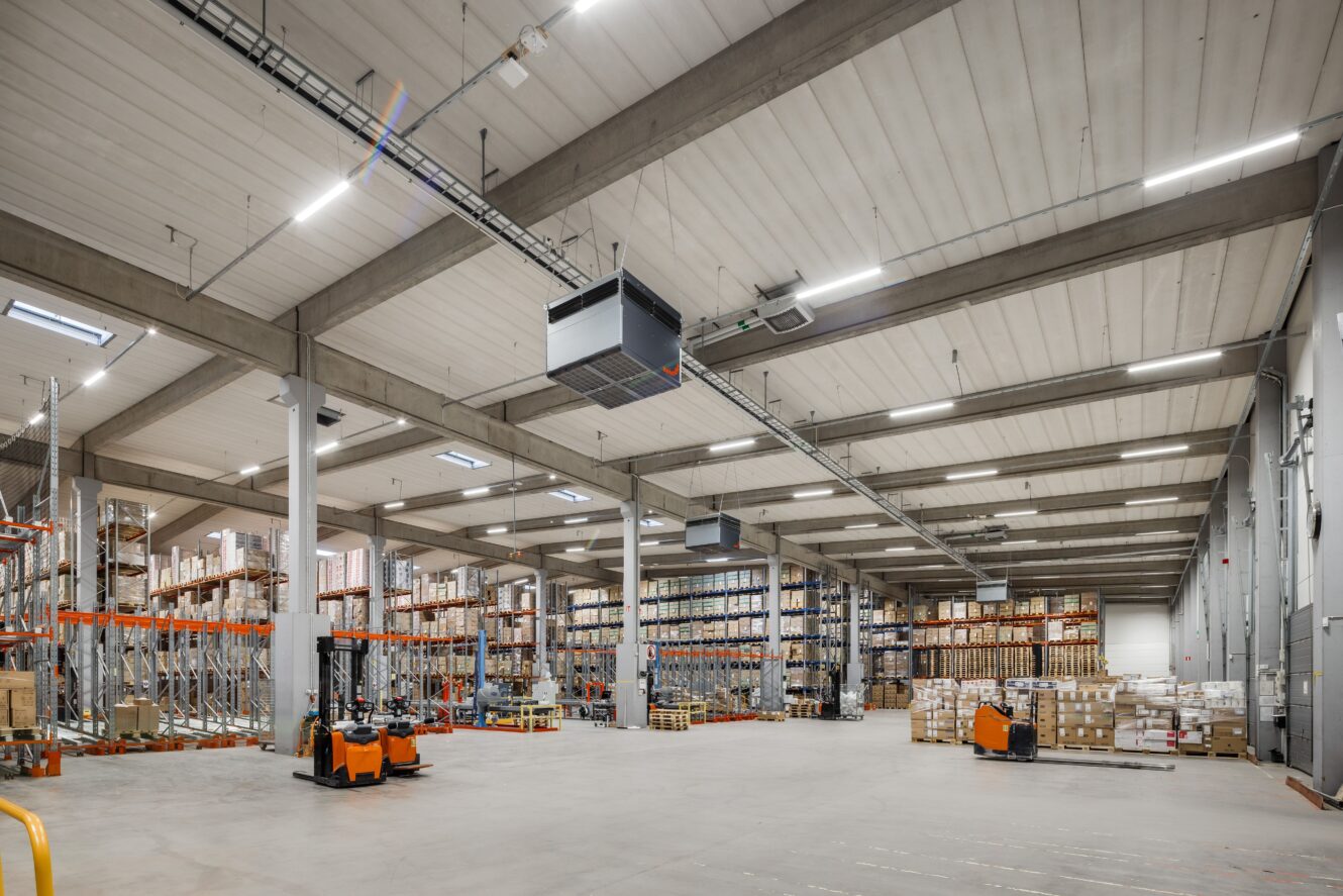 Stor lagerlokal med högt i tak, hyllor fyllda med lådor, industriell belysning och flera pallyftare på ett betonggolv.