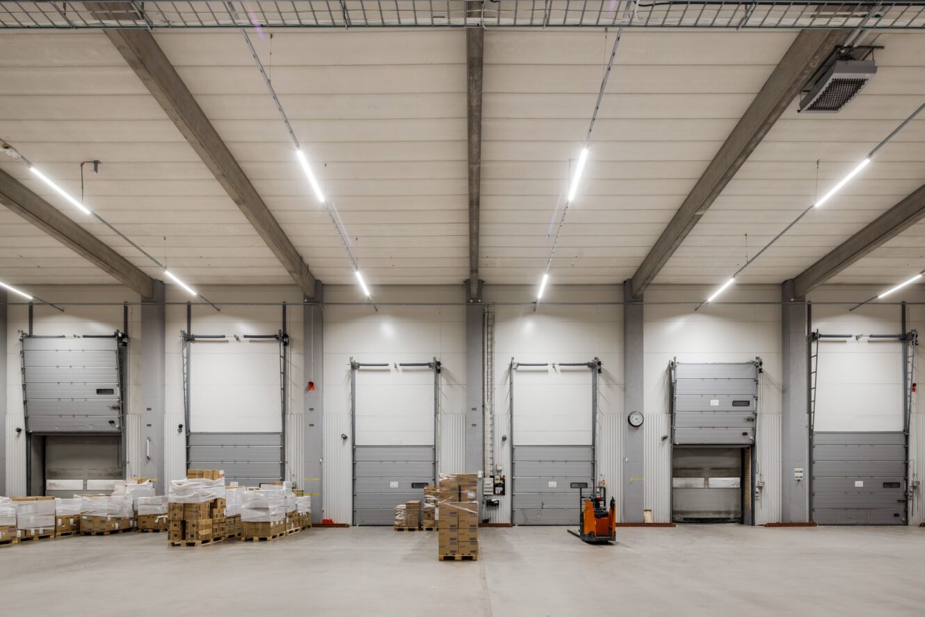 Interiör från en stor lagerlokal med lastbryggor, pallar med lådor och en orange pallbock placerad nära mitten. Området är väl upplyst och organiserat.