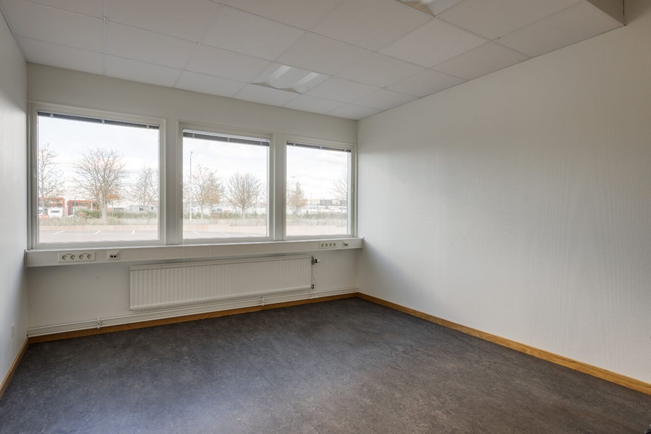 Ett tomt rum med stora fönster, vita väggar, en radiator under fönstren och mörkt golv. Naturligt ljus kommer in från utsidan och eluttag finns längs väggarna.