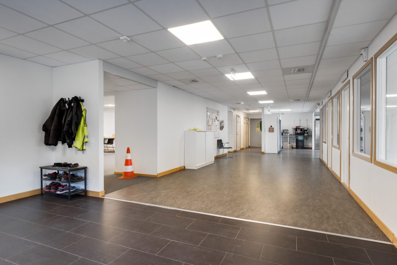 En ren, modern hall med vita väggar, klädkrokar, skohylla, en trafikkon och kontorsutrustning som syns i angränsande rum.