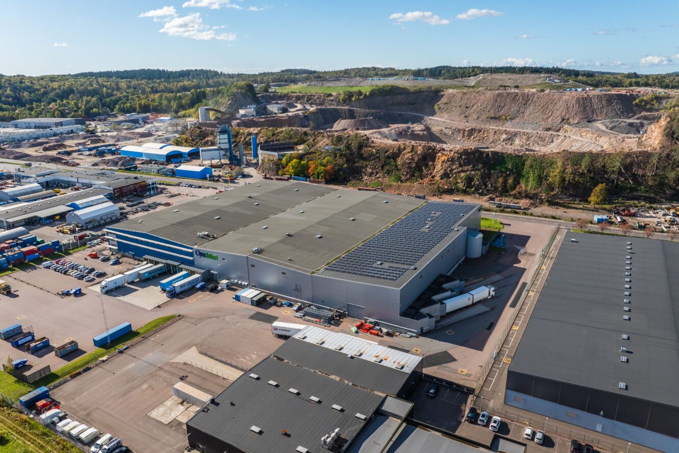 Flygfoto över ett industrikomplex med lagerlokaler, lastbilar och containrar, beläget nära ett stenbrott och omgivet av grönska under en klarblå himmel.