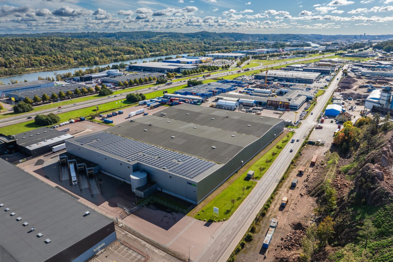 Flygfoto över ett industriområde med stora lagerbyggnader, vägar, grönska och en flod i bakgrunden under en delvis molnig himmel.