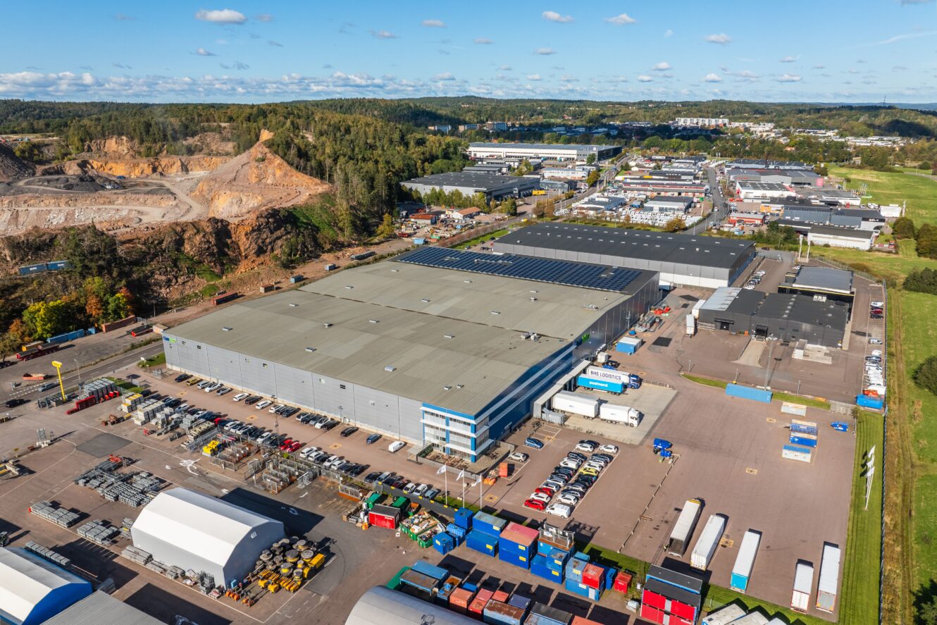 Flygfoto över ett industriområde med stora lagerlokaler, parkerade lastbilar, containrar och omgivande grönska under en delvis molnig himmel.