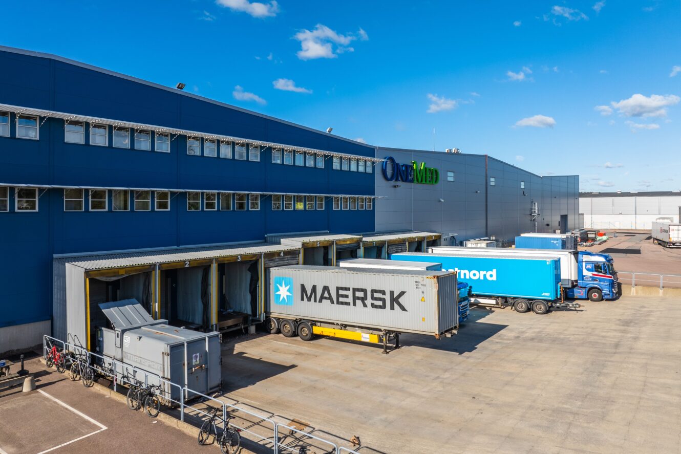 Lastbilar backade upp till lastbryggor vid ett stort blått och grått industriellt lager under en klar himmel.