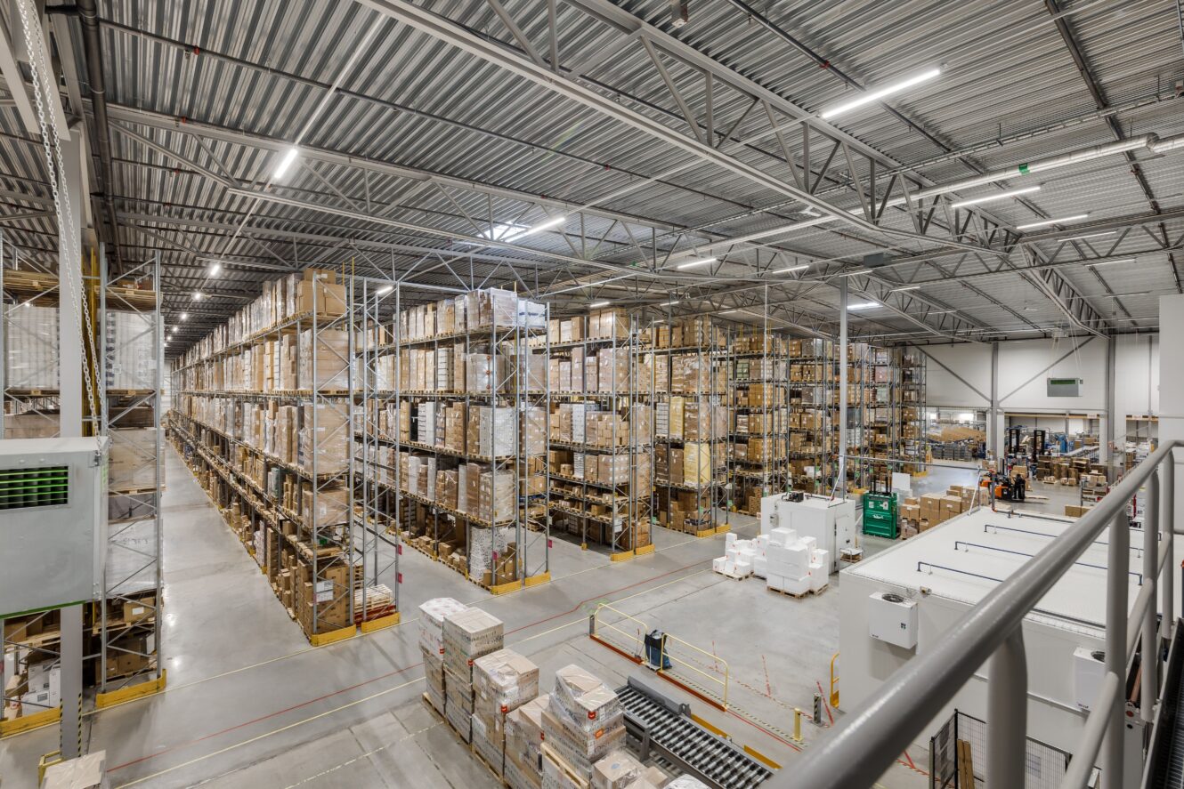 Stort lager med höga hyllor staplade med lådor och pallar, breda gångar och industriell belysning, sett från en upphöjd plattform.