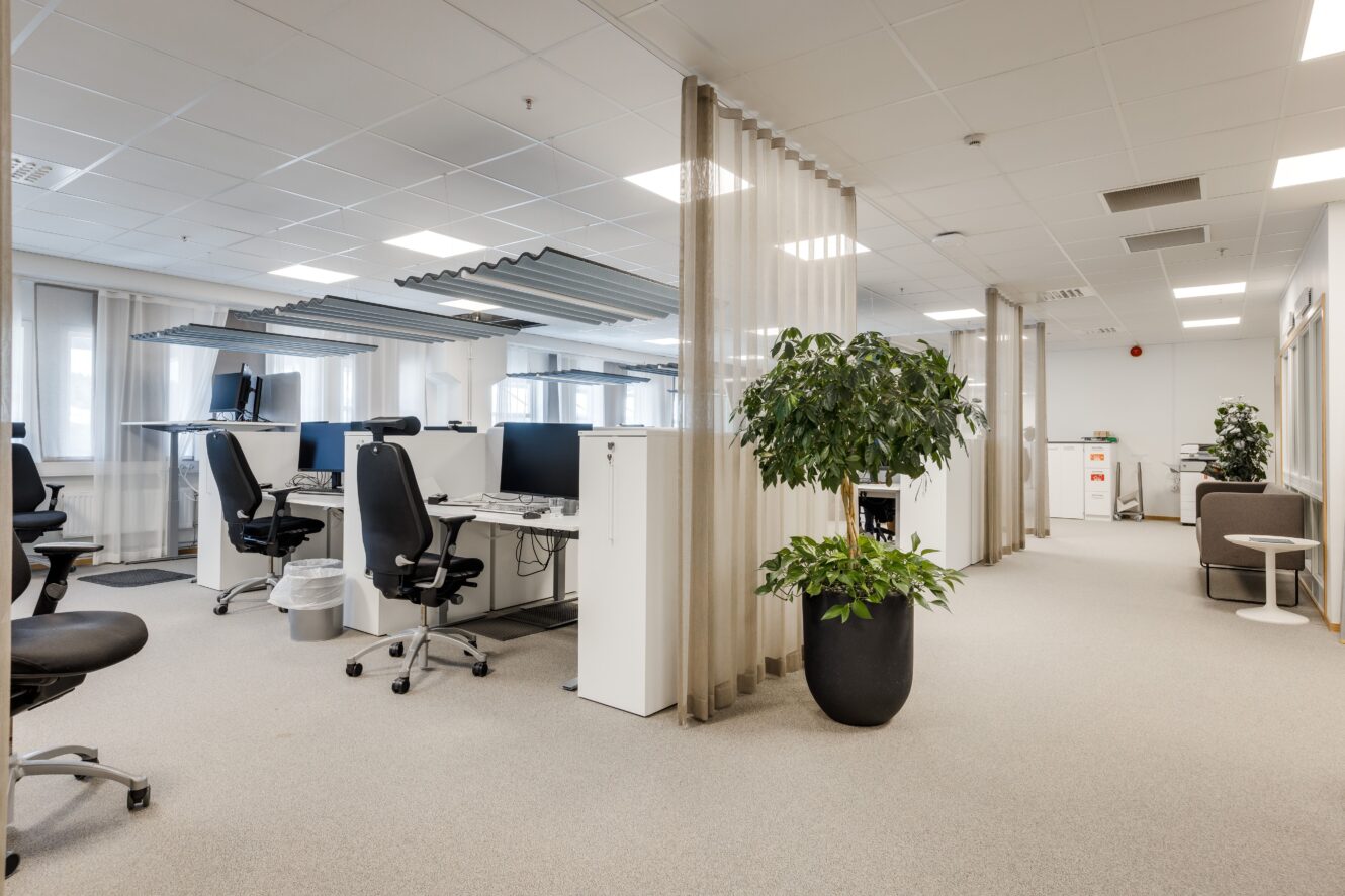 Moderna kontorslokaler med arbetsstationer, datorer, stolar, en stor krukväxt, ljusa mattor och glasväggar under starka taklampor.