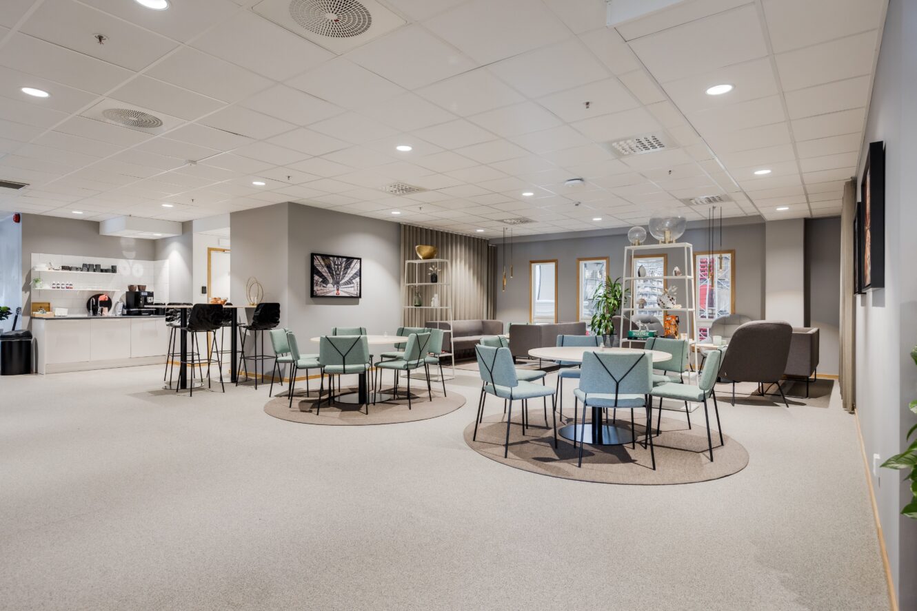 Modern kontorslounge med runda bord, pastellgröna stolar, höga stolar nära ett pentry, växter och neutral inredning under starka taklampor.