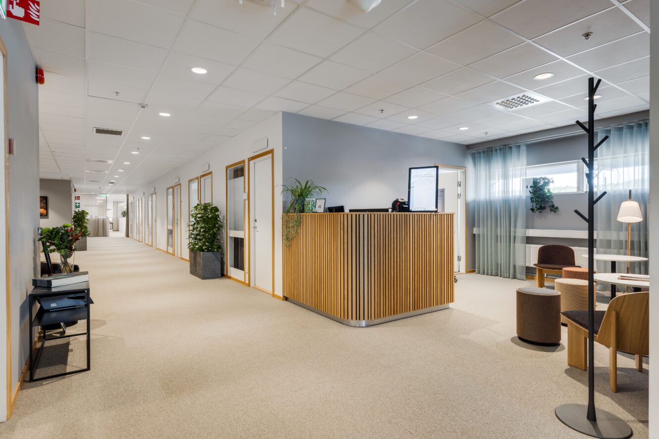 Modern kontorskorridor med receptionsdisk i trä, krukväxter, sittgrupp och flera dörrar som leder till enskilda kontor.