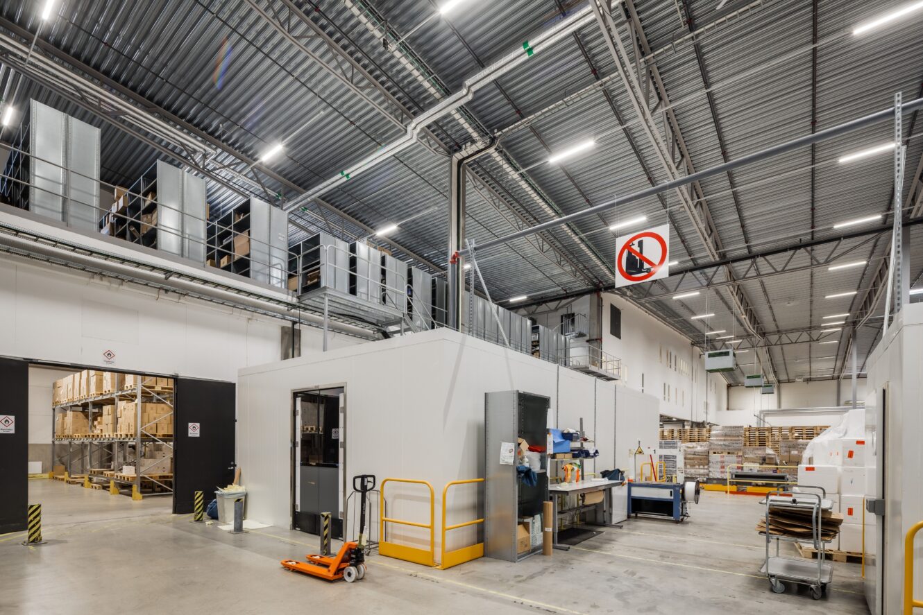 Interiörvy av en modern lagerlokal med industriell belysning, lagerhyllor, pallar och hanteringsutrustning; skylt om rökförbud synlig ovanför.