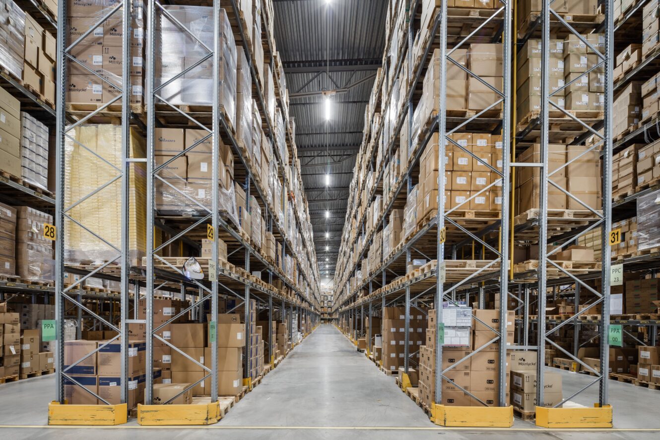 Stor lagerlokal med höga hyllor staplade med kartonger på båda sidor om en bred mittgång under stark takbelysning.