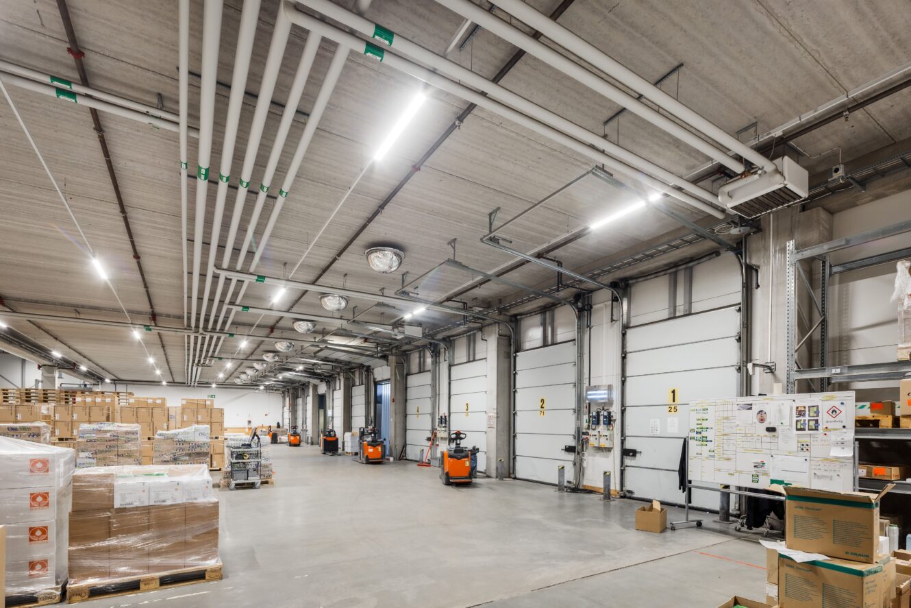 Stor lagerlokal med högt i tak, staplade lådor på pallar, orange pallbockar, vita dörrar till lastkajer samt rör och belysning ovanför.