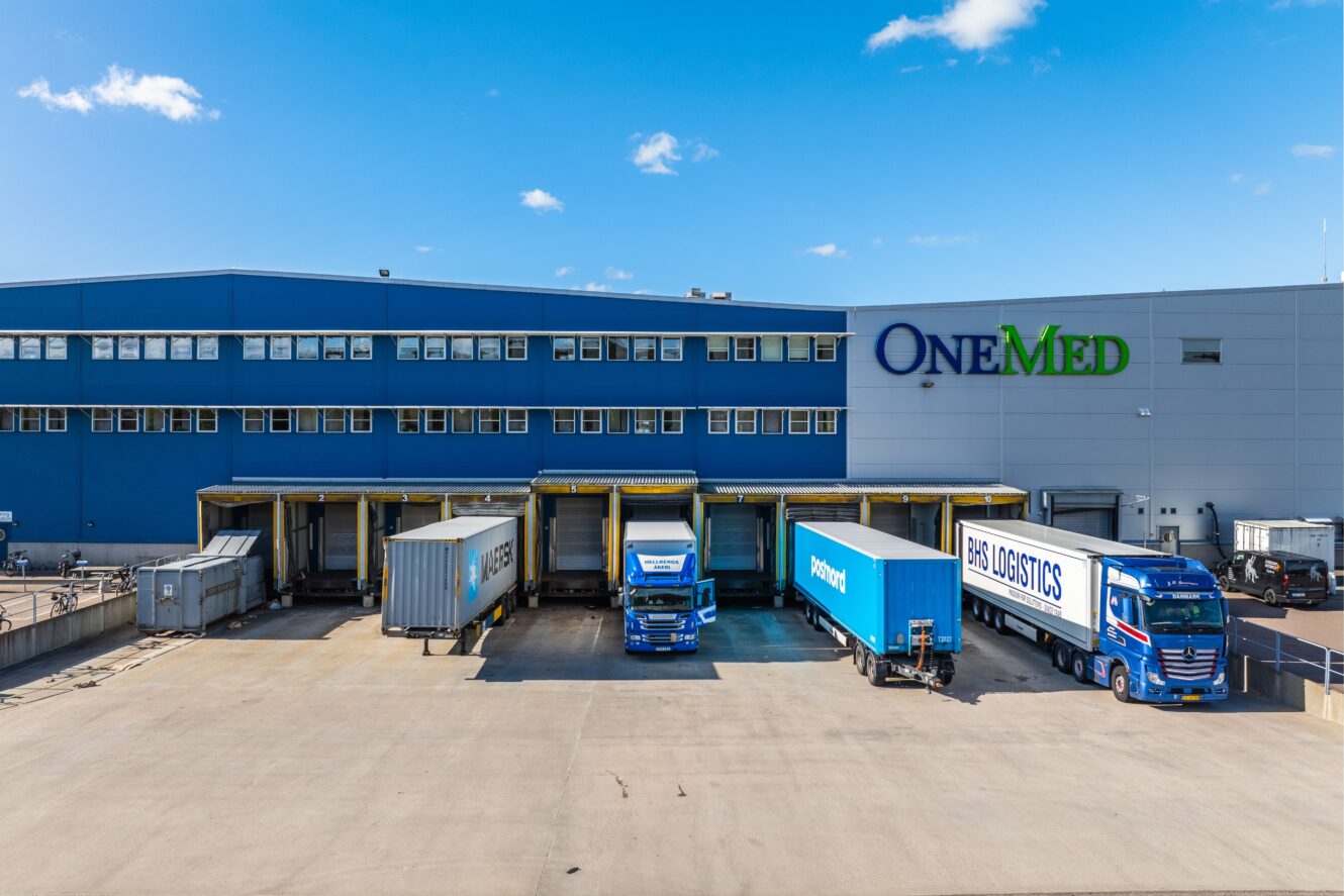 En stor lagerlokal märkt OneMed med flera lastbryggor, tre lastbilar parkerade vid kajer och klarblå himmel ovanför.
