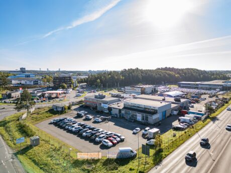Flygfoto över ett affärsområde med flera byggnader, parkeringsplatser fulla av bilar och närliggande vägar med fordon i rörelse under en ljus himmel.