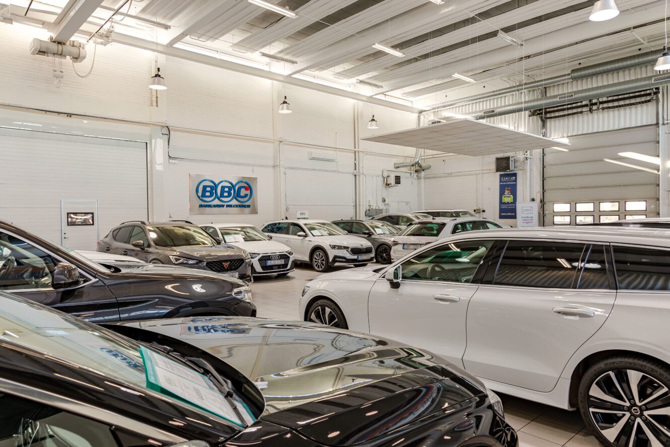 En bilhall visar flera parkerade fordon under stark takbelysning, med en BBC-skylt på bakväggen och stora garageportar synliga.