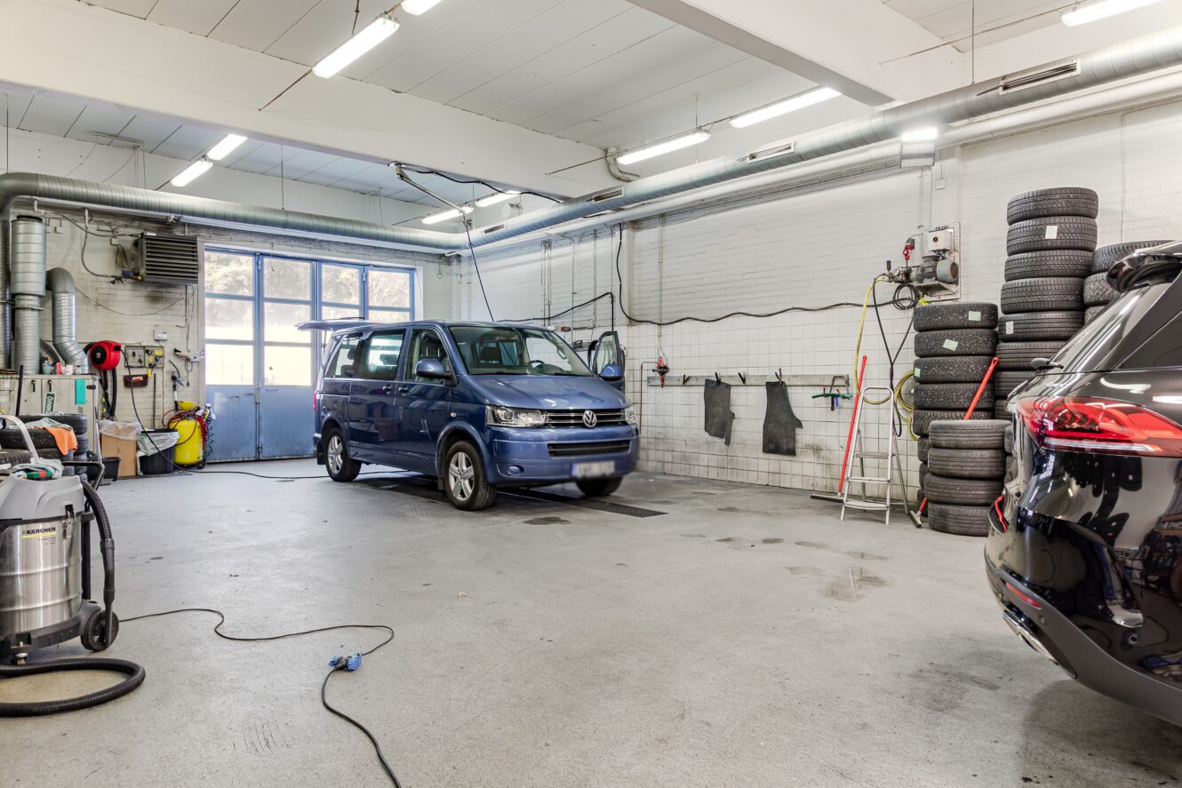 Interiör av en bilverkstad med en blå skåpbil parkerad inuti, staplade däck mot väggen, olika verktyg och utrustning synlig i hela utrymmet.